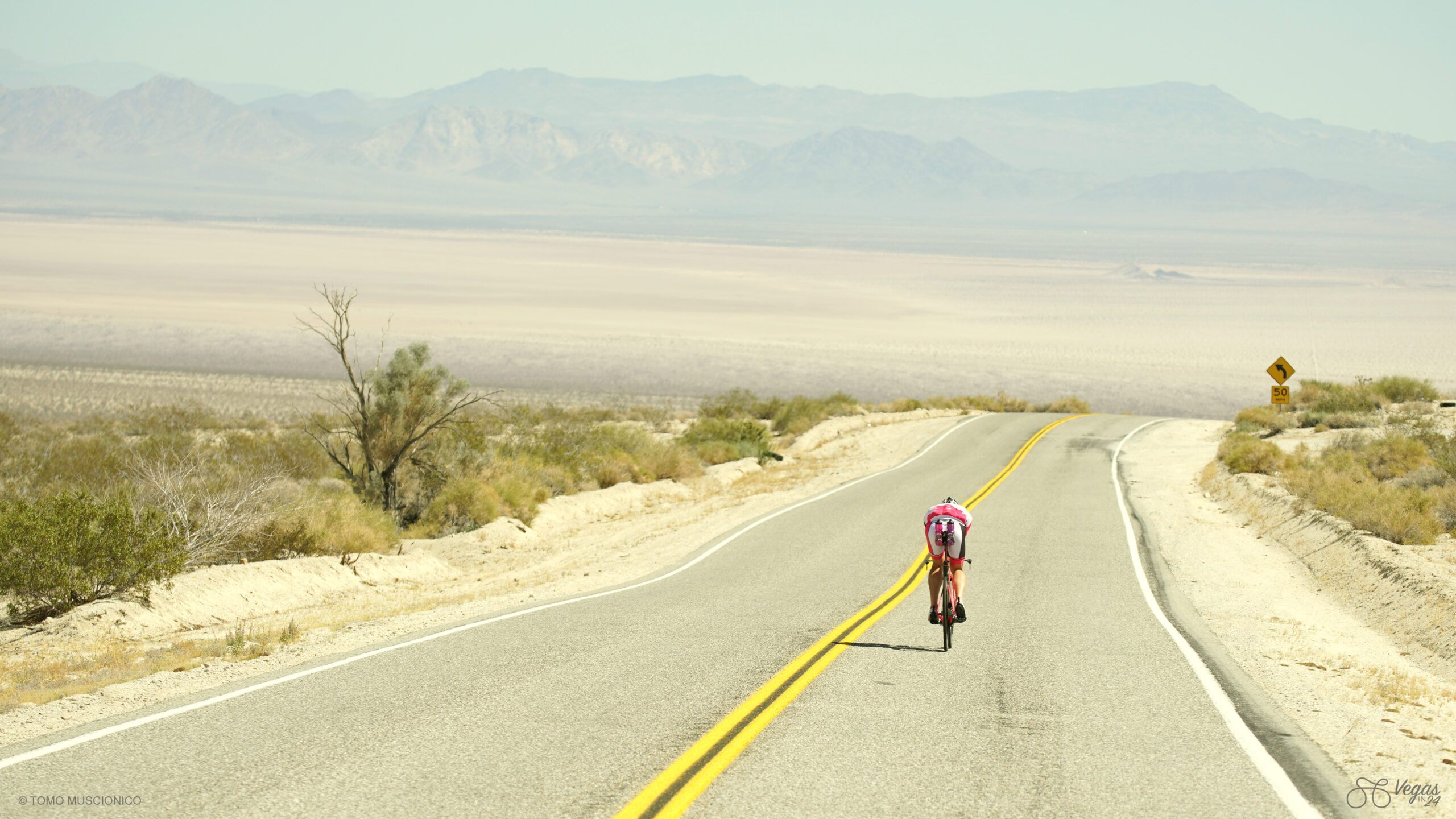 Andy Funk descends in aero position into the empty wilderness of the Mojave desert, captured on April 2016 during the original Vegas In 24 cycling challenge. A vast, arid desert landscape is visible with distant mountains beneath a hazy sky. Shrubs and sparse vegetation line the roadside.