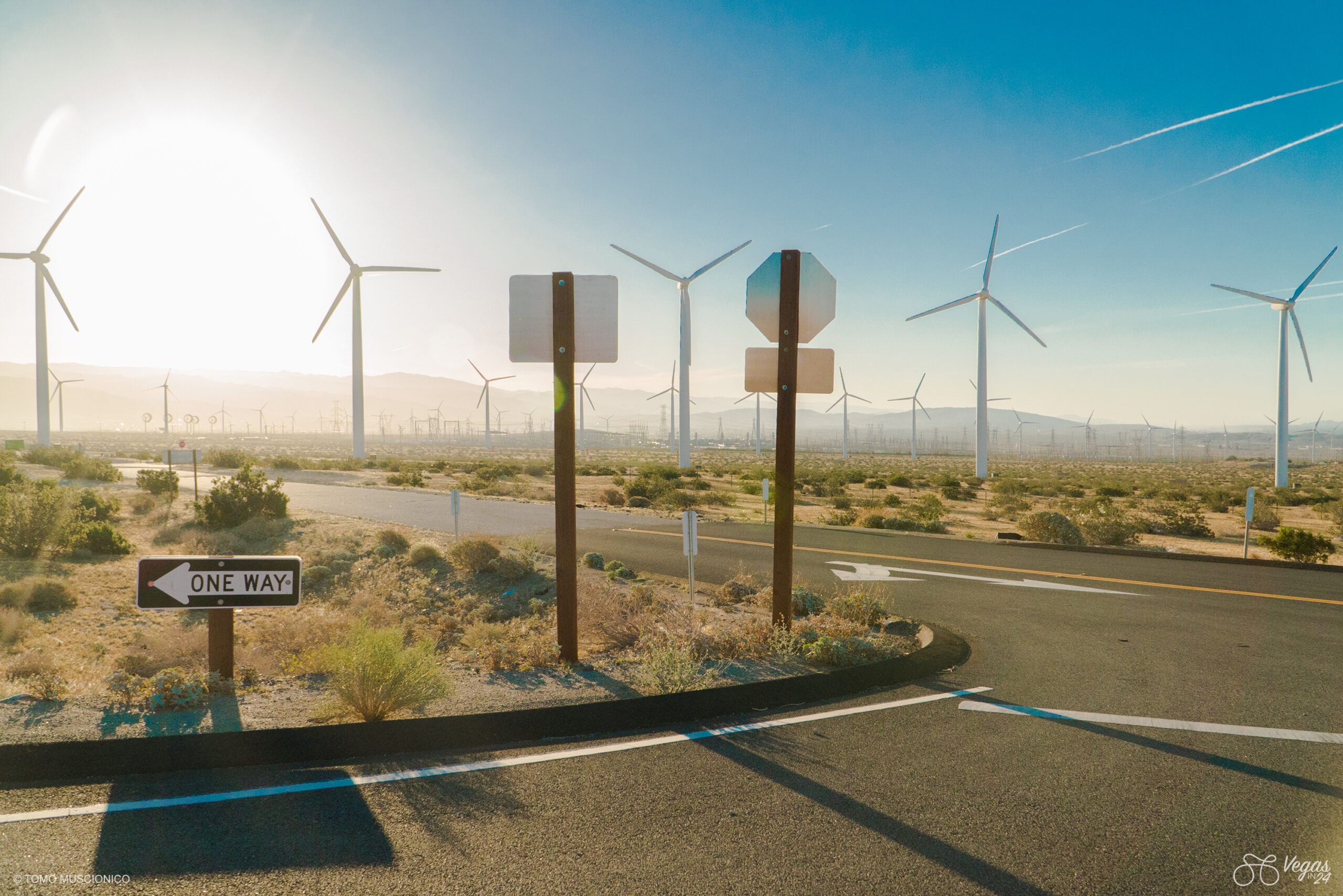 A road with a One Way sign curves through a desert landscape filled with wind turbines near the Palm Springs, California basin, on a windy day - captured during the early-morning hours during the inaugural Vegas In 24 ultra-cycling challenge in 2016. The sun shines brightly, casting long shadows, while mountains rise in the background.