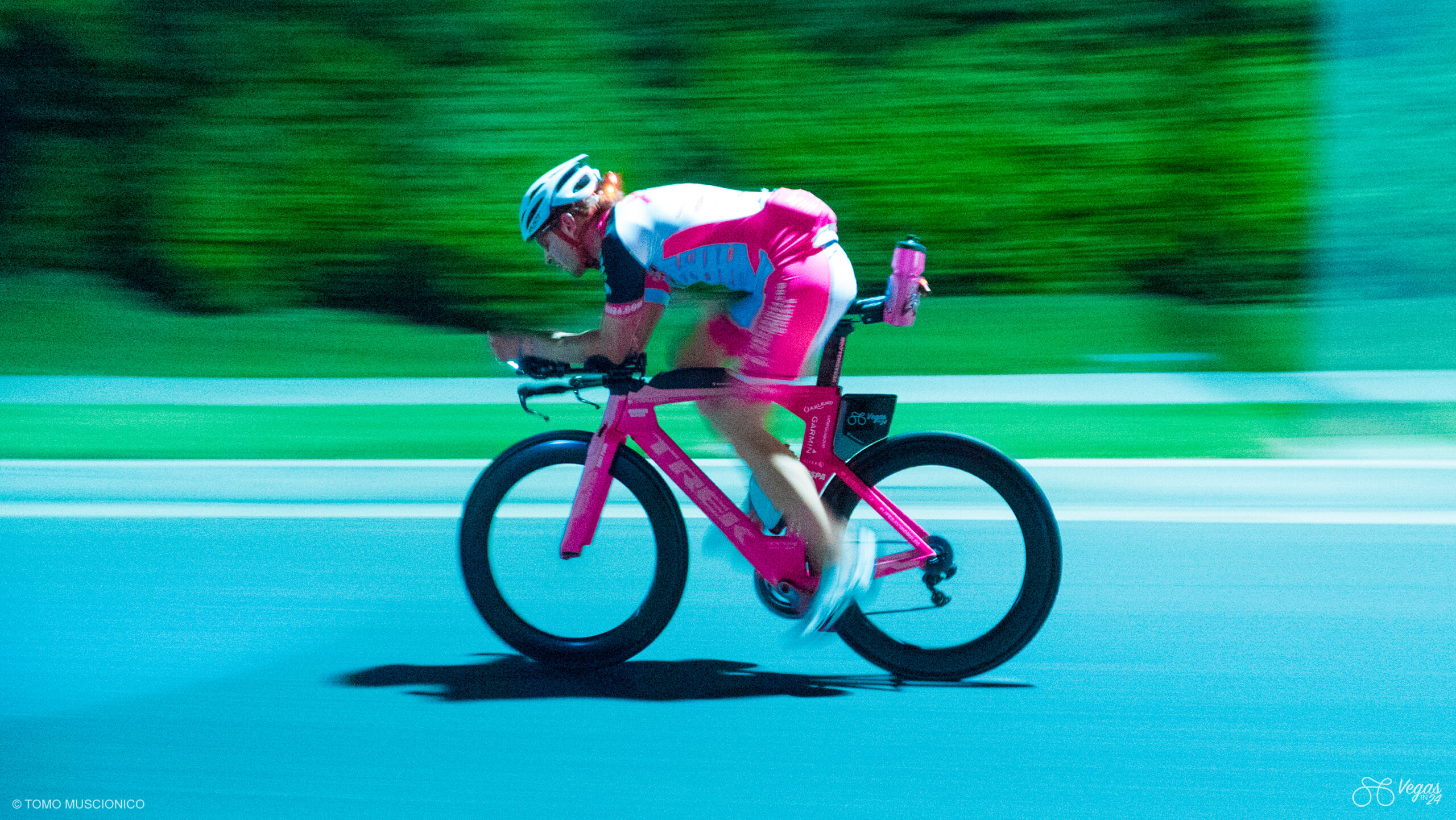 Andy Funk, Ho'Oamu Chief & Founder, in a pink and white outfit rides a his bike at high speed in the middle of the night during the 2016 Vegas In 24 cycling challenge, tilted forward in an aerodynamic position.