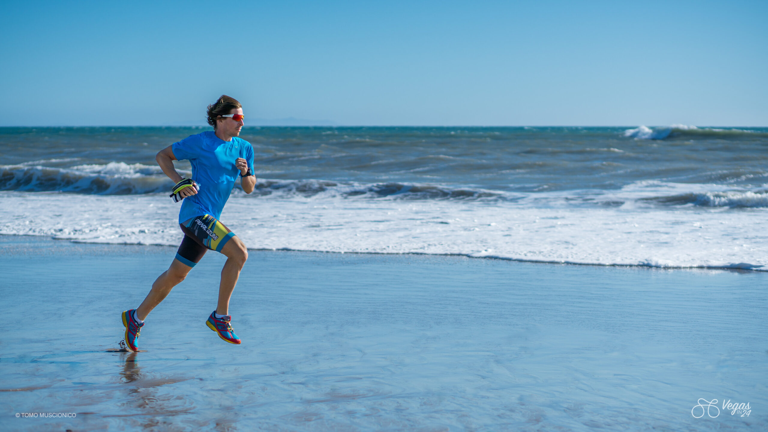 Serial entrepreneur and Ho'Omau founder Andy Funk, wearing a blue shirt, shorts, and sunglasses, runs along the shoreline of Santa Monica, waves in the background under a clear blue sky during one of his final cross-training runs before taking on the inaugural Vegas In 24 cycling challenge.