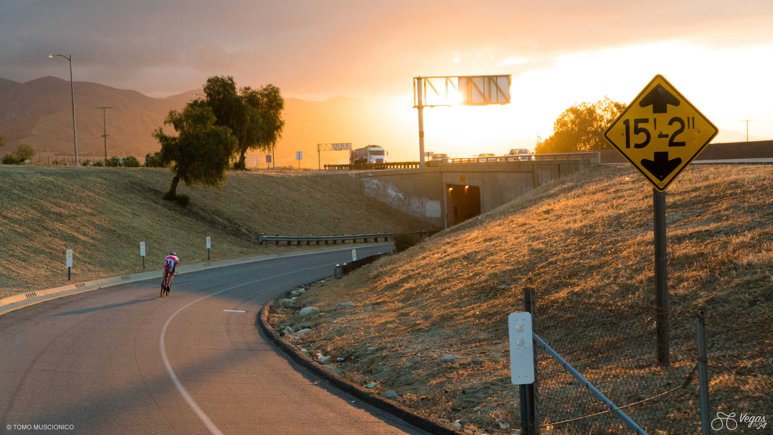 After 5 hours in the saddle, Andy Funk, founder of Ho'Omau, rides into the first moments of sunrise on April 23, 2016, pedaling along a curved road near an overpass in Beaumont, California prior to reaching his first rest stop on his 365-mile journey. A yellow 15-2 clearance sign on the right and cars are trailing in the background - captured during the first-ever Vegas In 24 ultra-cycling challenge.