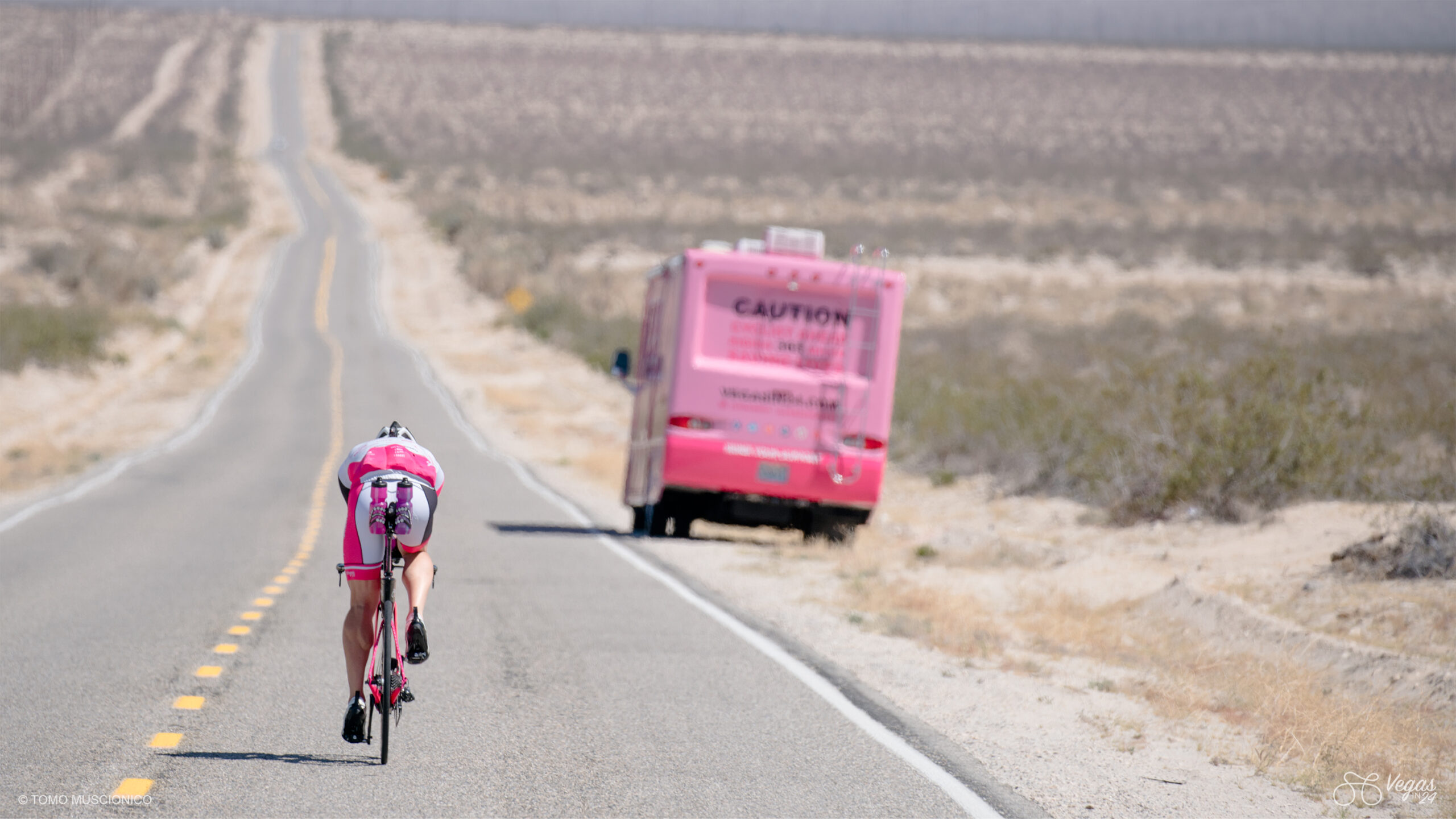 Ho'Omau Founder Andy Funk rides along an empty desert road towards Amboy, California during the original 2016 Vegas In 24 cycling challenge, about to pass a parked pink RV. The dry landscape features sparse vegetation and distant mountains beneath a clear sky.