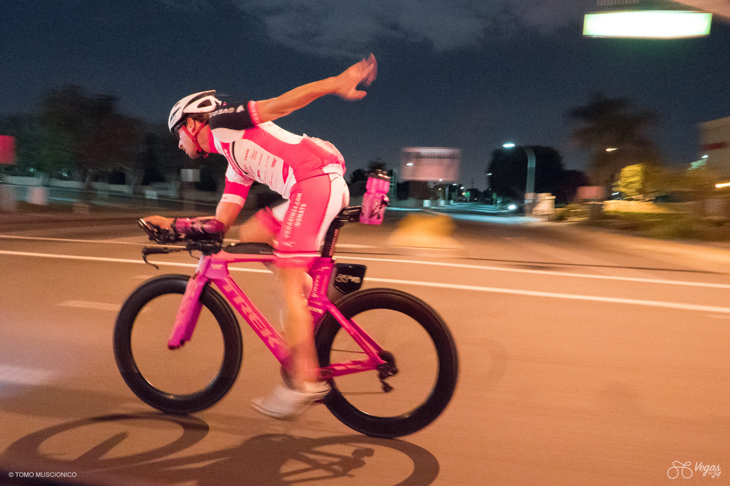 Ho'Omau founder and entrepreneur Andy Funk rides his pink bike on a street in the middle of the night, extending one arm backward to stretch out his shoulders and back - captured during the inaugural Vegas In 24 ultra-cycling challenge in 2016.