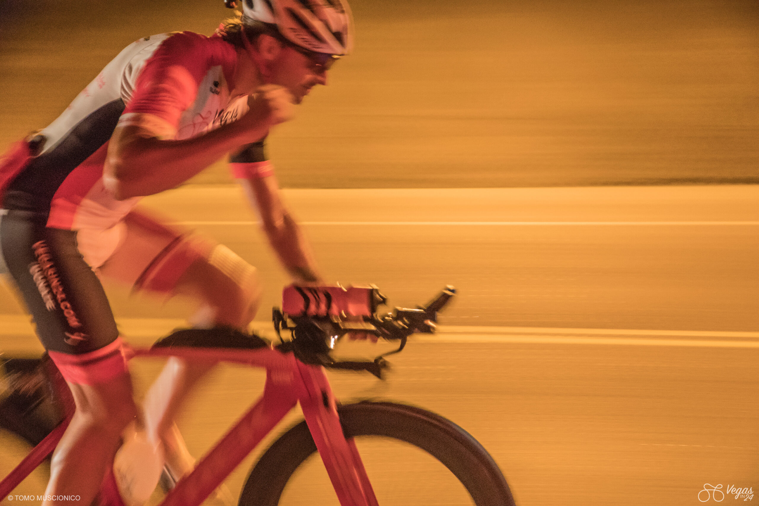 With Las Vegas visible in the distance ahead, Andy Funk, Ho'Omau Chief, raises his fist in celebration that the finish line is within reach. Captured during the early evening hours of the inaugural Vegas In 24 cycling challenge in 2016. Motion blur highlights the speed of the ride under warm, orange-toned lighting.