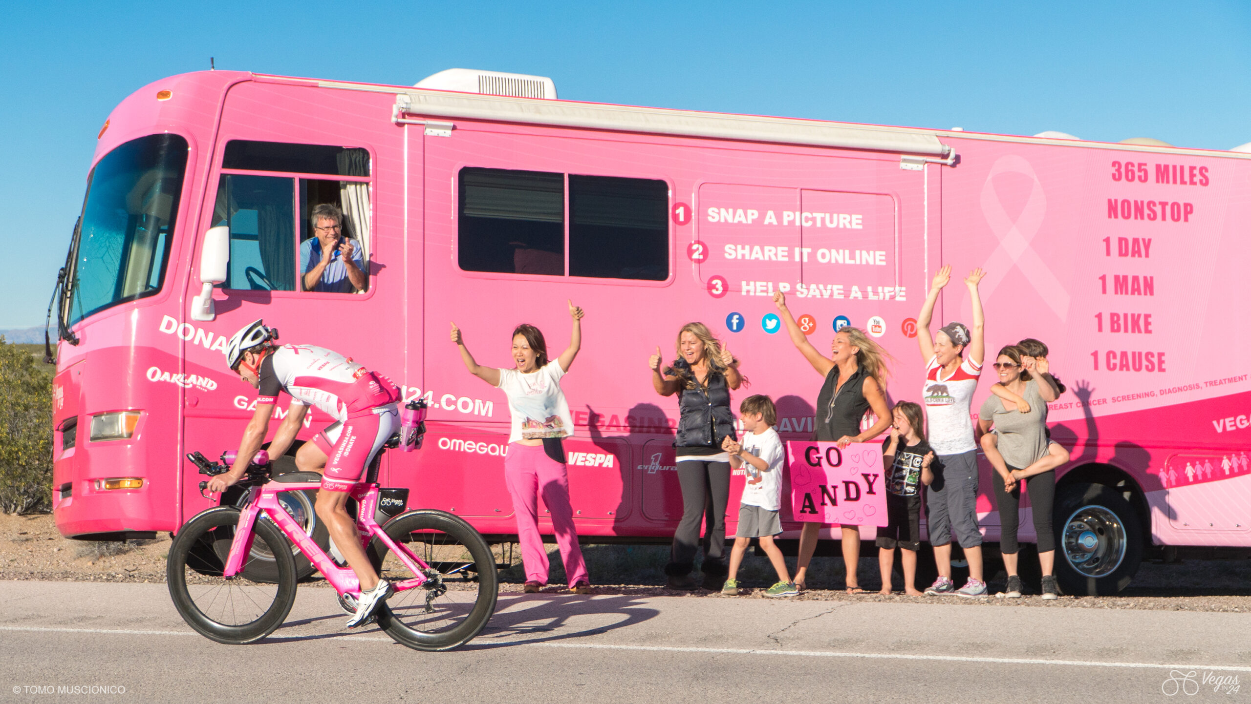 Halfway of the final long climb to Searchlight, Nevada, Andy Funk, Founder of Ho'Omau Endurance, is on the final leg of the inaugural 365-mile Vegas In 24 ultra-cycling challenge. A cheering group, including his wife Dr. Kristi Funk and children, hold signs and wave enthusiastically from the roadside. The RV is decorated in matching pink with encouraging messages.