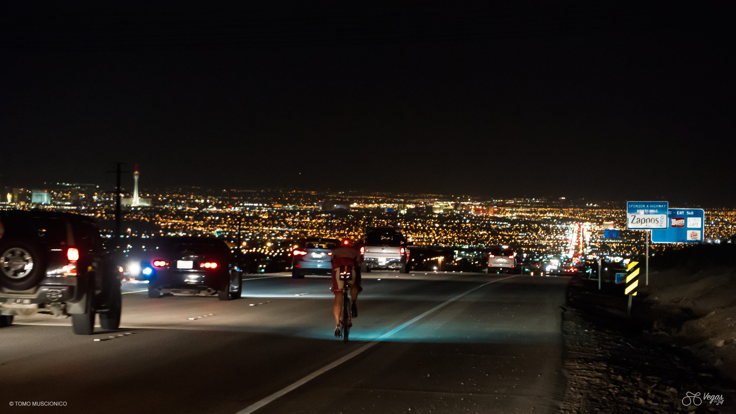 In the final hour of the inaugural 2016 Vegas In 24 ultra-cycling challenge, Andy Funk, Chief Ho'Omau, crests the final short climb towards the finish line on a busy, illuminated road at night, surrounded by cars and the glowing Las Vegas cityscape in the distance. Road signs appear on the right side of the image.