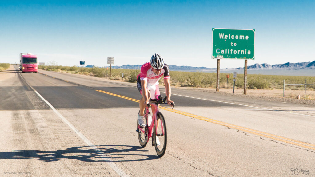 Ho'Omau athlete and founder Andy Funk, in pink and white gear, rides on a desert highway, with the green "Welcome to California" sign shown on the opposite side of the street. Captured mid-day in April 2016 during the original 365-mile Vegas In 24 ultra-cycling challenge. Desert landscape and a pink RV appear in the background under a clear blue sky.