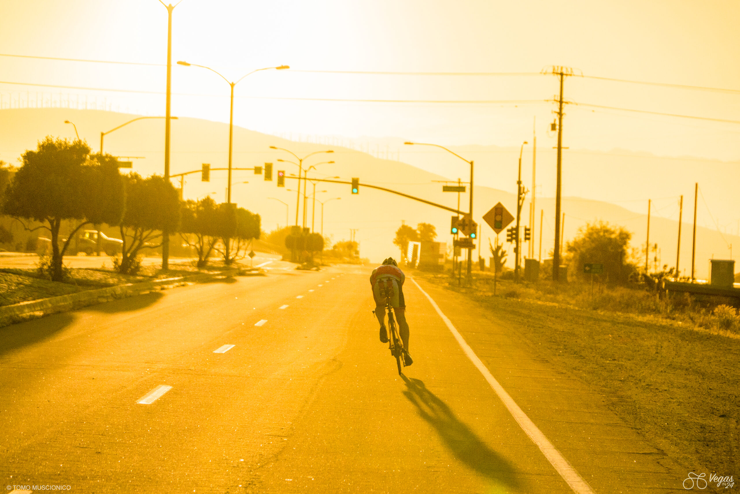 Andy Funk, Ho'Omau Founder & CEO, rides alone on an empty road at sunrise, as golden sunlight casts long shadows. His Trek Speed Concept is tilted heavily towards the left, counter-acting the strong cross winds present at the time. Traffic lights, street lamps, trees, and distant hills fade into the warm, hazy background. Captured during the early-morning hours of the original 2016 Vegas In 24 ultra-cycling challenge.