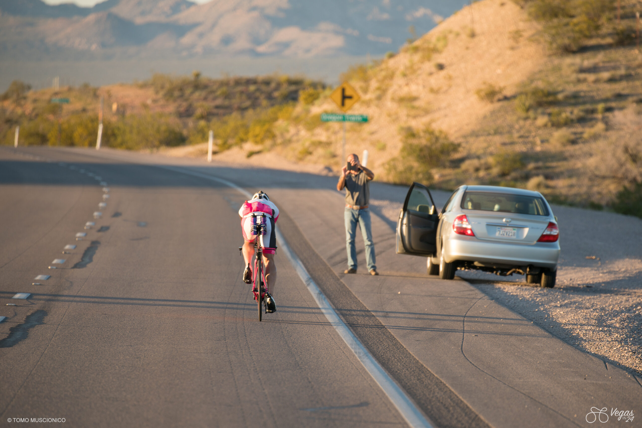 With only a few hours left to the finish line, Andy Funk, endurance athlete and Ho'Omau founder, embarks on the final stretch of the inaugural Vegas In 24 ultra-cycling challenge in April 2016. A person beside a parked car captures the moment with his phone against the backdrop of mountains and desert landscape.