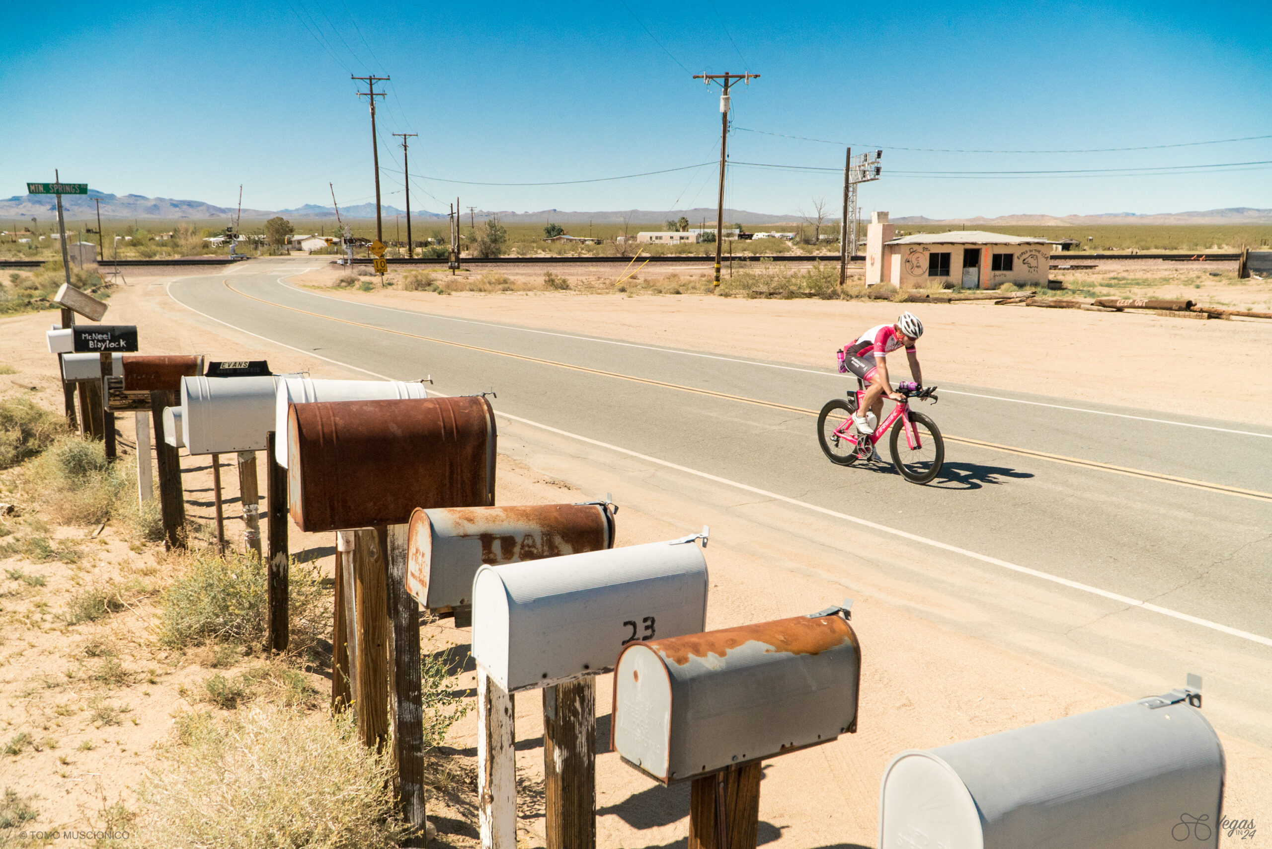 Endurance athlete and entrepreneur Andy Funk rides along a desert road, passing old mailboxes amid dry terrain, sparse vegetation, power lines, and distant mountains under a clear blue sky. Captured during the mid-day hours of the original Vegas In 24 ultra-cycling challenge, heading towards the California Nevada border.