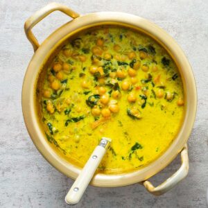 A pot of golden-yellow saffron stew with chickpeas and greens sits on a gray surface in this Race-Ready Kitchen scene, surrounded by fresh herbs, bread, a small pitcher of vegan cream, and salt and pepper shakers.
