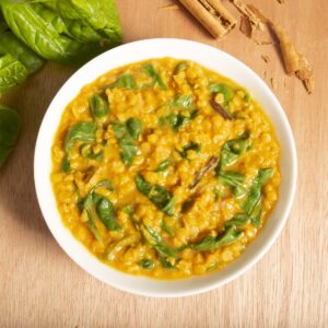 A bowl of yellow lentil curry with spinach sits on a wooden surface, surrounded by cinnamon sticks and spinach leaves.