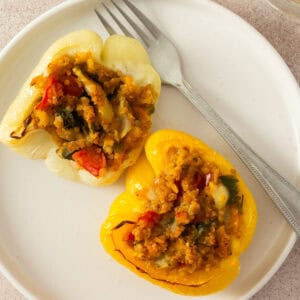 A white plate holds two yellow bell peppers stuffed with a hearty filling of vegetables and grains, showcasing a Race-Ready Kitchen creation. A fork rests nearby, and a glass of water with lemon sits at the top edge of the image.