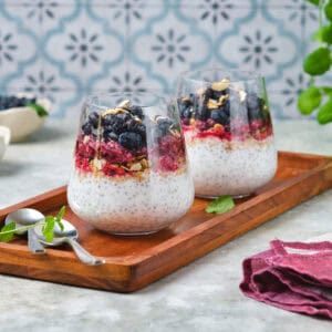 Two glasses of chia pudding topped with berries and seeds sit on a wooden tray in this Race-Ready Kitchen scene. Garnished with mint, the setup includes a purple napkin and patterned tile wall.