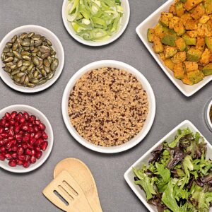 Top-down view of a quinoa bowl in a Race-Ready Kitchen, surrounded by small dishes of pumpkin seeds, pomegranate seeds, leafy greens, and a wooden spatula on a beige surface.
