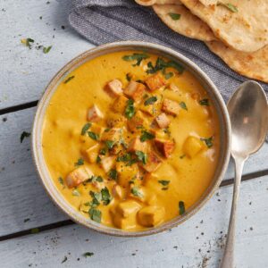 A bowl of vegan tikka masala curry topped with chopped herbs and grilled tofu sits on a blue wooden surface next to sliced naan and a spoon.