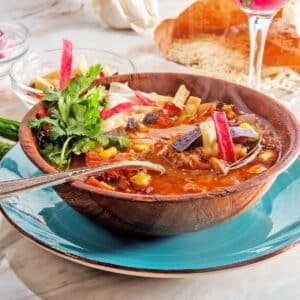 A wooden bowl of colorful Aztec Biome-Booster Soup, garnished with fresh herbs and vegetables, sits on a blue plate.