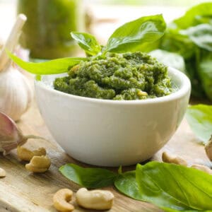 A small white bowl of green pesto is surrounded by fresh basil leaves, garlic cloves, and cashew nuts on a wooden surface.