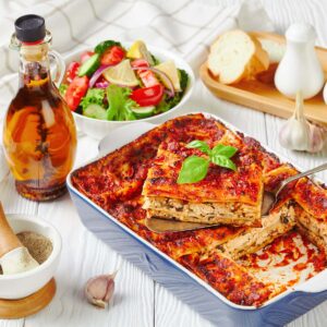 A baked lasagna in a blue dish is shown with a serving on a spatula, surrounded by salad, bread, tea, garlic, and salt and pepper shakers.