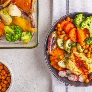 A colorful bowl filled with roasted vegetables, broccoli, chickpeas, radishes, and hummus sits next to a baking dish of veggies—perfect for a Race-Ready Kitchen. A small bowl of chickpeas is also visible.