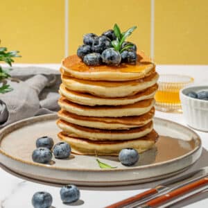 A stack of pancakes topped with blueberries and a mint sprig sits on a plate with extra blueberries around it. Utensils and a small bowl of blueberries complete this Race-Ready Kitchen scene.