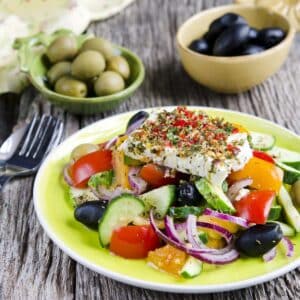 A colorful Mediterranean salad with sliced cucumbers, tomatoes, onions, peppers, olives, and herbed vegan feta cheese is served on a green plate in a Race-Ready Kitchen set on a rustic wooden table with bowls of olives in the background.