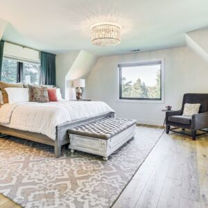 Spacious bedroom with large bed, armchair, wooden furniture, patterned rug, chandelier, and a window overlooking trees—perfect for luxury athlete accommodations in Lake Arrowhead during the Ho'Omau Altitude Haus training camp. Light-colored walls and hardwood floors complete the serene retreat.