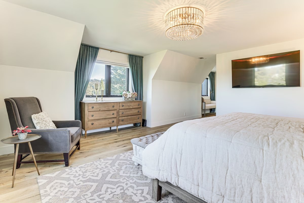 Bright, spacious bedroom in luxury athlete accommodations Lake Arrowhead during the Ho'Omau Altitude Haus training camp, featuring a large bed, armchair, side table, modern chandelier, dresser under blue-curtained window, wall-mounted TV, and light wood flooring with a patterned rug.