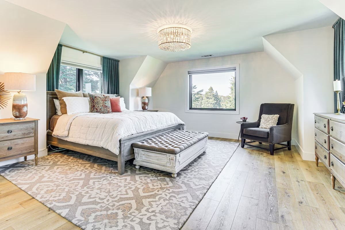 Spacious bedroom with large bed, armchair, wooden furniture, patterned rug, chandelier, and a window overlooking trees—perfect for luxury athlete accommodations in Lake Arrowhead during the Ho'Omau Altitude Haus training camp. Light-colored walls and hardwood floors complete the serene retreat.