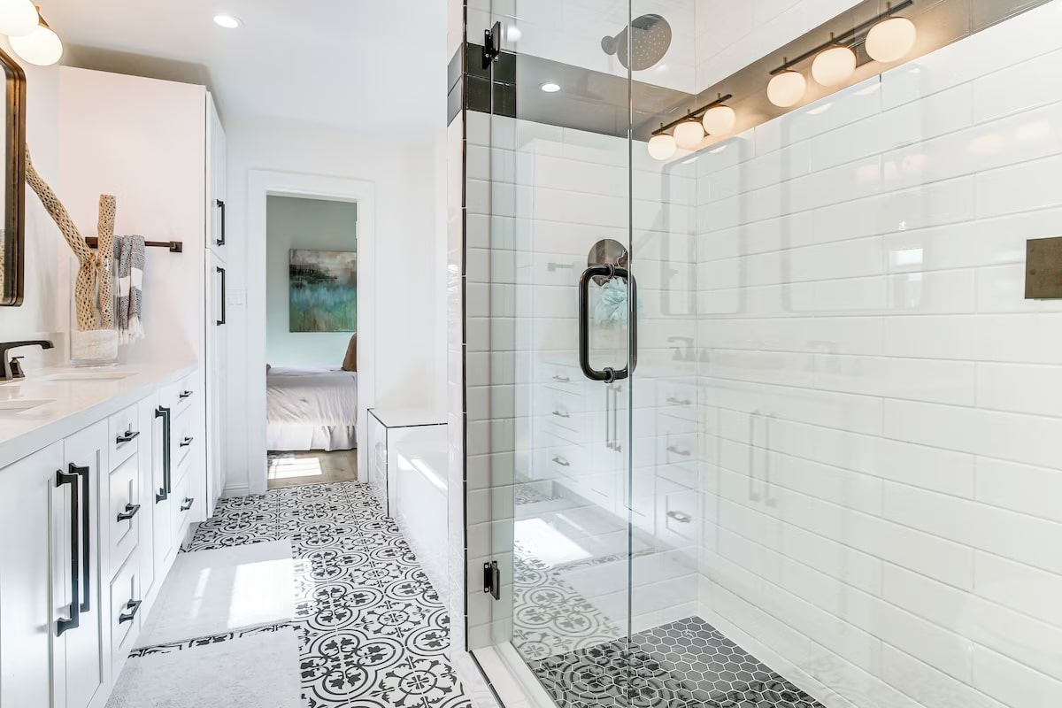 Modern bathroom with patterned tile floor, white subway tile shower with glass door, double sinks and white cabinets—designed for luxury athlete accommodations in Lake Arrowhead during the Ho'Omau Altitude Haus training camp. An open doorway leads to a bedroom in the background.