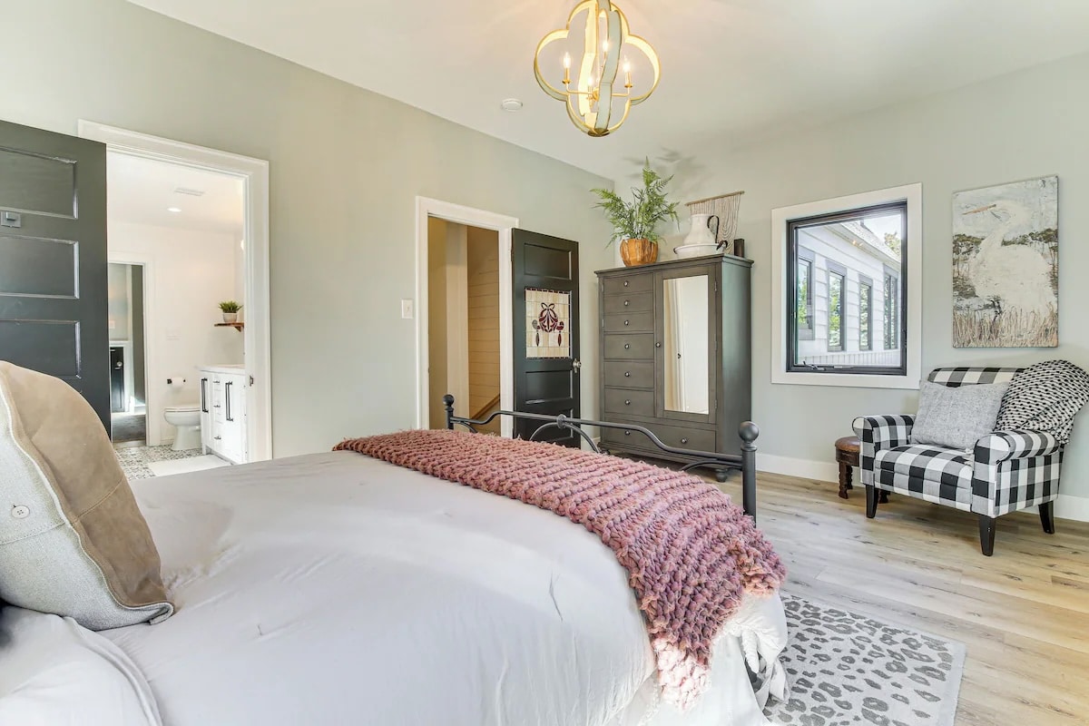 Bright bedroom with a made bed, pink knitted throw, gray dresser with plants, and art on the walls—exuding the comfort and style found in luxury athlete accommodations in Lake Arrowhead during the Ho'Omau Altitude Haus training camp. Adjacent bathroom visible through open door.