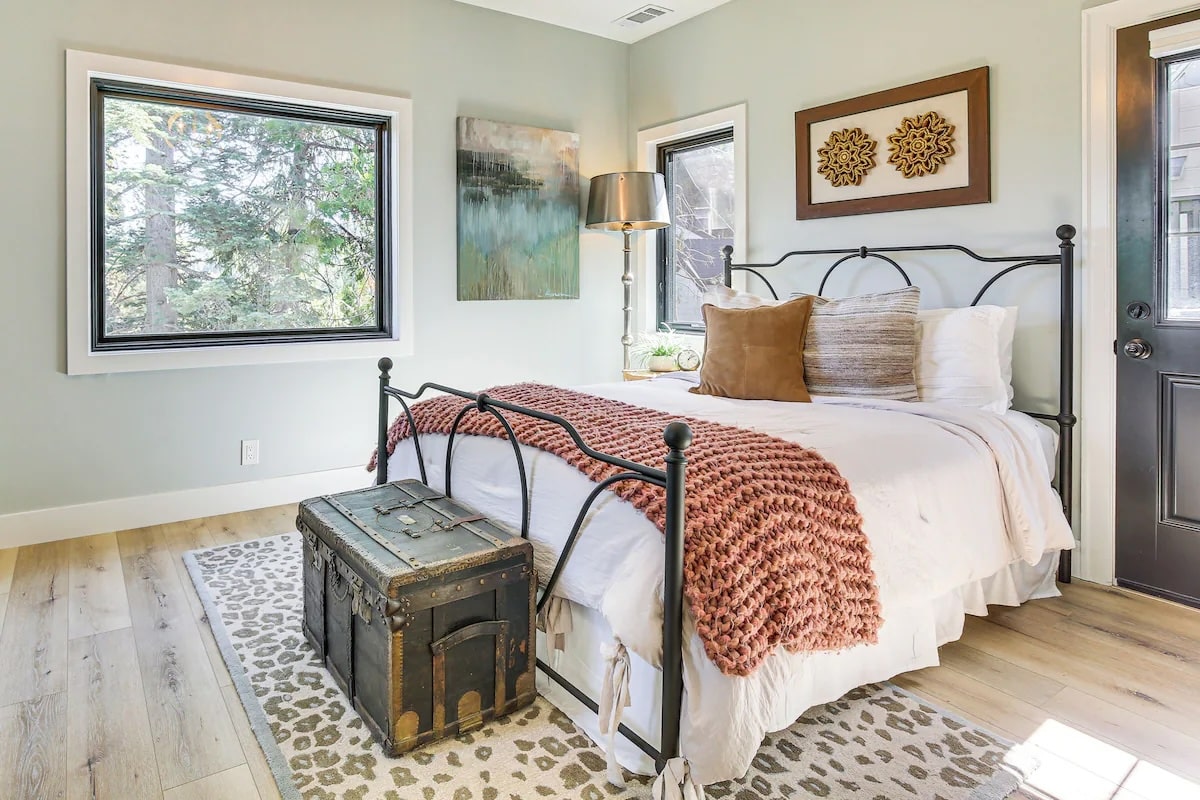 Bright bedroom with a black metal bed, white bedding, brown and pink throw blanket, decorative pillows, vintage trunk, artwork, a large window, and light wood flooring with a patterned rug—ideal for luxury athlete accommodations in Lake Arrowhead during the Ho'Omau Altitude Haus training camp.