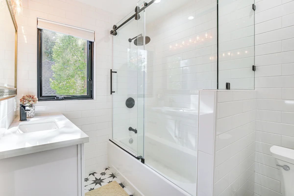 Modern bathroom with white subway tile, a glass-enclosed shower-tub combo, black fixtures, and a white vanity—evoking the style of luxury athlete accommodations in Lake Arrowhead during the Ho'Omau Altitude Haus training camp. Patterned floor tiles and floral accents complete the serene retreat.
