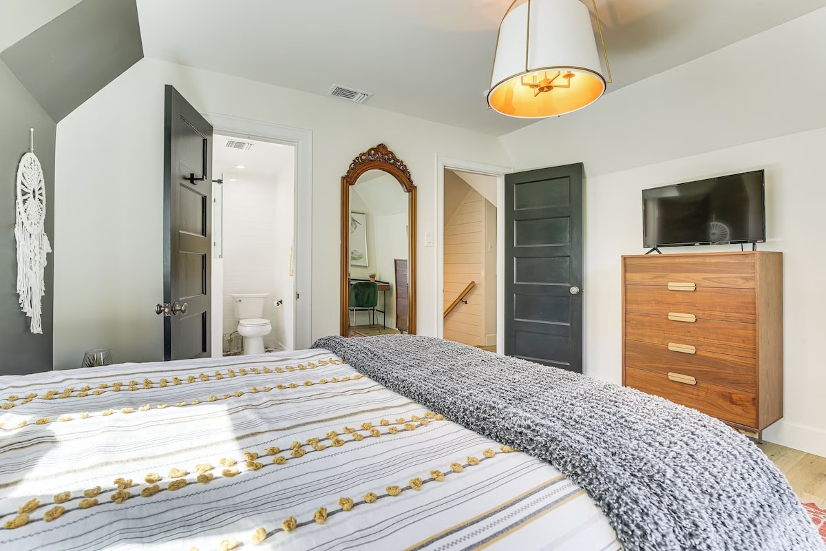 Bright bedroom in luxury athlete accommodations, featuring a striped bedspread, gray knit blanket, wooden dresser with TV, tall mirror, and dark doors connecting to bathroom and hallway—perfect for your stay in Lake Arrowhead during the Ho'Omau Altitude Haus training camp.