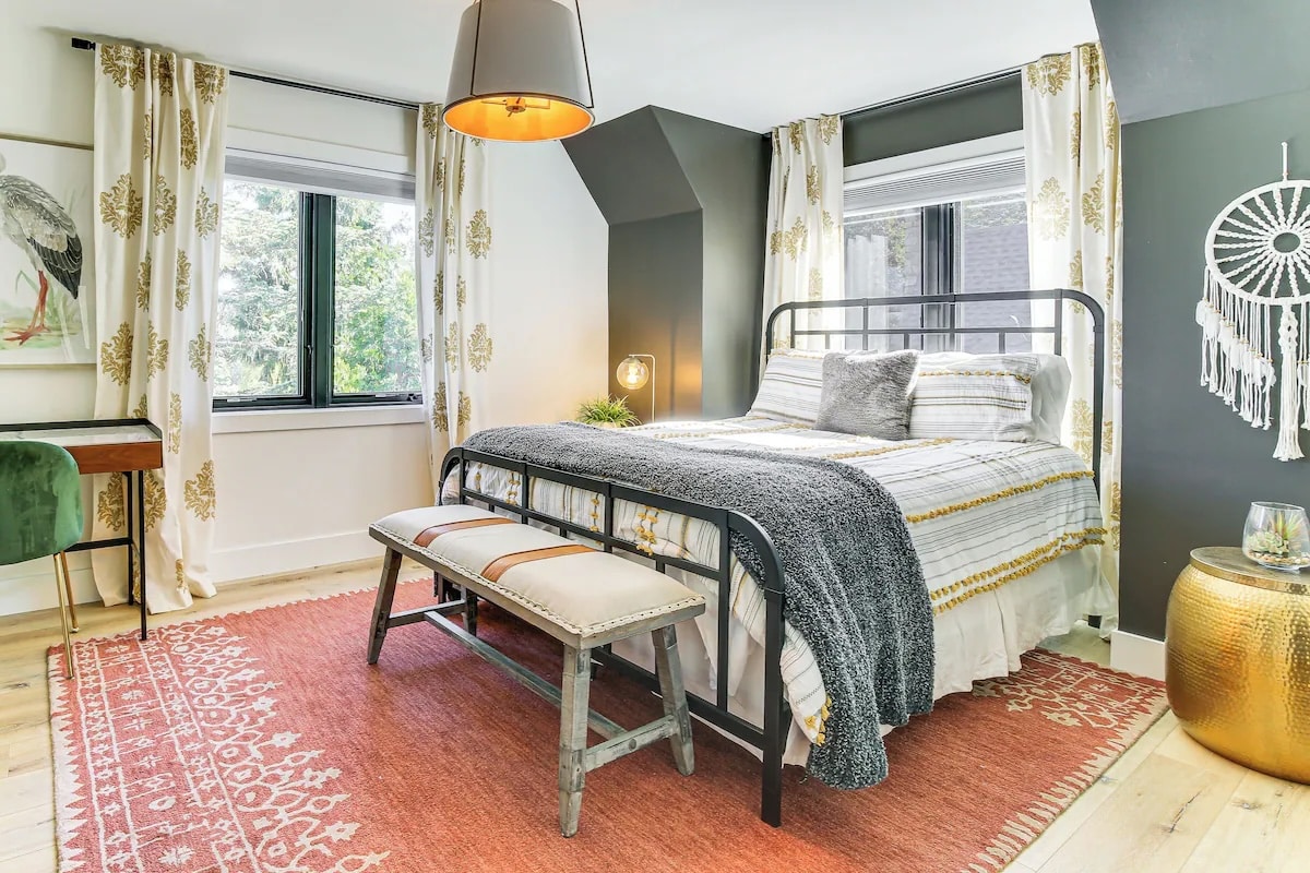 A bright bedroom with a metal bed, striped bedding, gray throw, and decorative pillows. Perfect for luxury athlete accommodations in Lake Arrowhead during the Ho'Omau Altitude Haus training camp, it features a coral rug, bench, lush plants, artwork, and abundant natural light for a cozy feel.