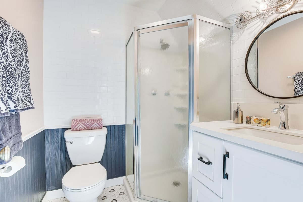 A modern bathroom in luxury athlete accommodations at Lake Arrowhead during the Ho'Omau Altitude Haus training camp, featuring a glass shower, white vanity, round mirror, rolled towels, white tile with dark wainscoting, and stylish patterned floors.