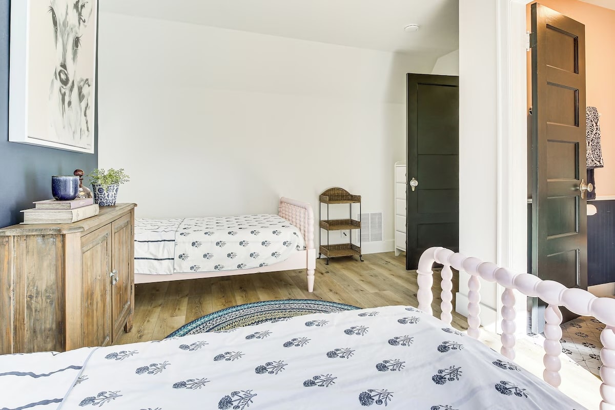 Bright bedroom with two twin beds featuring white bedding with black floral prints, a wooden floor, a wooden cabinet with decor, and a small wicker shelf; perfect for luxury athlete accommodations in Lake Arrowhead during the Ho'Omau Altitude Haus training camp. One door opens to a lit hallway.