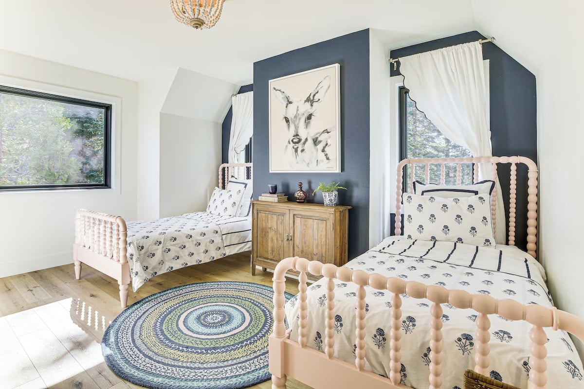 Bright, airy bedroom with two light wood twin beds, patterned bedding, a round braided rug, wooden dresser, and large windows with white curtains—perfect for luxury athlete accommodations in Lake Arrowhead during the Ho'Omau Altitude Haus training camp. Deer artwork adorns the blue accent wall.