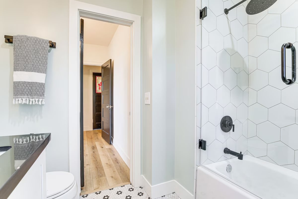 Modern bathroom in luxury athlete accommodations during the Ho'Omau Altitude Haus training camp, featuring a white hexagon tile shower, black fixtures, towel on the wall, white walls, black countertop, and an open door leading to a hallway with wood flooring in Lake Arrowhead.