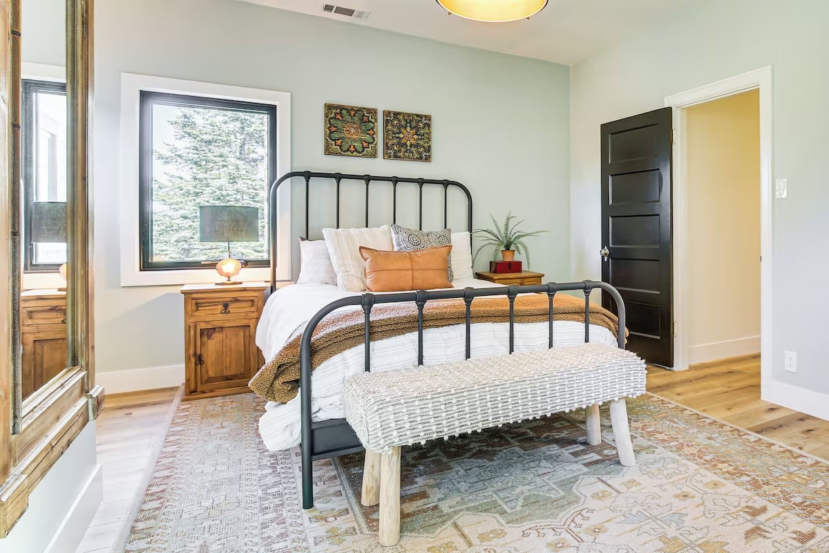 Bright bedroom with a metal bed frame, layered bedding, and a woven bench at the foot. Luxury athlete accommodations in Lake Arrowhead during the Ho'Omau Altitude Haus training camp include wood nightstands with lamps, light walls adorned with art, and a patterned rug over light wood floors.