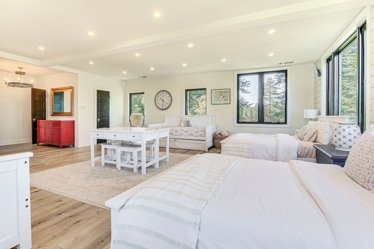 A spacious, bright bedroom with large windows, two twin beds, a double bed, a central desk, red dresser near the entry, and light wood flooring. Enjoy neutral decor and nature views—ideal for luxury athlete accommodations in Lake Arrowhead during the Ho'Omau Altitude Haus training camp.