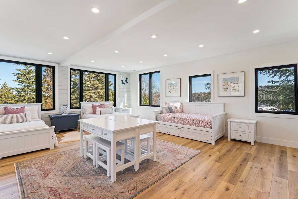 Bright, spacious room with large windows, two daybeds, a white table with benches, and wooden floors—perfect for luxury athlete accommodations in Lake Arrowhead during the Ho'Omau Altitude Haus training camp. Framed art decorates the walls and trees are visible outside.