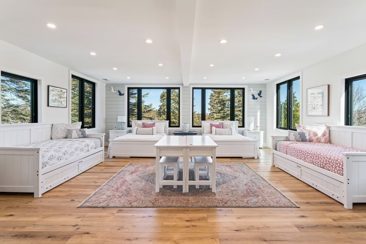 Bright, spacious room with large windows, three white daybeds with patterned bedding, a central table and chairs, light wood floors, and scenic views of trees—ideal for luxury athlete accommodations in Lake Arrowhead during the Ho'Omau Altitude Haus training camp.