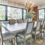 A bright dining room inspired by luxury athlete accommodations in Lake Arrowhead during the Ho'Omau Altitude Haus training camp, featuring a rustic wooden table, eight striped chairs, a modern gold chandelier, large windows with gold curtains, floral centerpiece, and nature-themed wallpaper.
