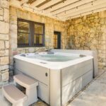 A modern hot tub with bubbling water sits on a concrete patio against a stone wall, offering luxury athlete accommodations in Lake Arrowhead during the Ho'Omau Altitude Haus training camp, near a black door, two windows, and string lights hanging from a white wooden ceiling.