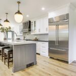Modern kitchen with white cabinets, stainless steel appliances, a large gray island with barstools, pendant lights, wooden floors, and a patterned backsplash. Large windows provide natural light in these luxury athlete accommodations at Lake Arrowhead during the Ho'Omau Altitude Haus training camp.