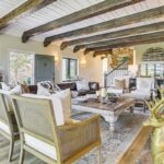 A cozy living room in luxury athlete accommodations in Lake Arrowhead during the Ho'Omau Altitude Haus training camp, with rustic wood beams, a stone fireplace, eclectic decor, a leather sofa, cane armchairs, and a gold reindeer sculpture on a patterned rug bathed in natural light.