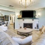 A cozy living room inspired by luxury athlete accommodations in Lake Arrowhead, featuring a curved gray sectional sofa, round coffee table, stone fireplace with green mantel, mounted TV, bookshelves, and a modern chandelier. Light pours in from the right.
