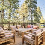 A cozy outdoor patio with cushioned wooden chairs around a table, two tall patio heaters, string lights overhead, a barbecue grill, and surrounded by tall pine trees—perfect for luxury athlete accommodations in Lake Arrowhead during the Ho'Omau Altitude Haus training camp.