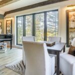 Bright room with large windows, a black grand piano, four white upholstered chairs around a dark wooden table, nature artwork on walls, light wood floors, an exposed wood beam, and a zebra-patterned rug evoke luxury athlete accommodations in Lake Arrowhead during the Ho'Omau Altitude Haus training camp.